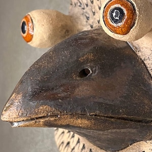 Pu&ograve; includere: Una scultura da parete in ceramica di una testa di uccello con un becco marrone e due grandi occhi con dettagli arancioni e neri. La testa dell'uccello &egrave; testurizzata con piccole linee in rilievo.