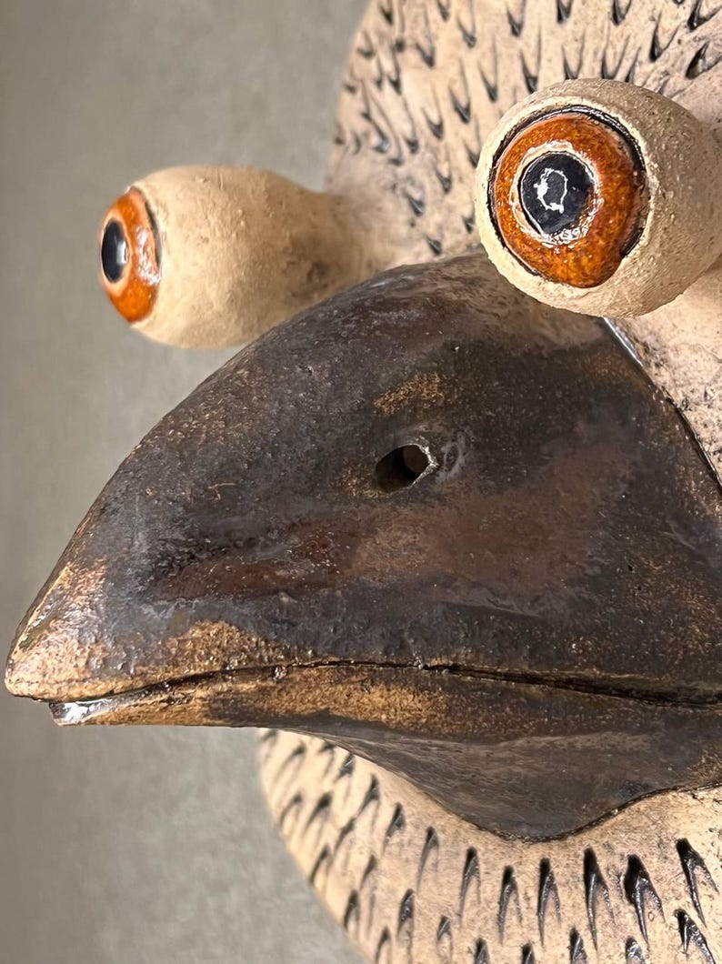 Pu&ograve; includere: Una scultura da parete in ceramica di una testa di uccello con un becco marrone e due grandi occhi con dettagli arancioni e neri. La testa dell'uccello &egrave; testurizzata con piccole linee in rilievo.