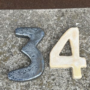 Op de afbeelding: Twee decoratieve cijfers, 3 en 4, worden getoond. Het cijfer 3 is een donker, getextureerd metaal met een verweerde uitstraling. Het cijfer 4 is lichtbeige van kleur met een glad oppervlak. De cijfers liggen op een getextureerd, grijs oppervlak.