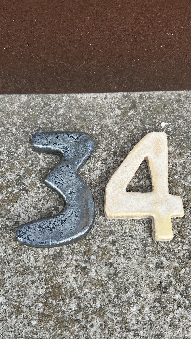 Op de afbeelding: Twee decoratieve cijfers, 3 en 4, worden getoond. Het cijfer 3 is een donker, getextureerd metaal met een verweerde uitstraling. Het cijfer 4 is lichtbeige van kleur met een glad oppervlak. De cijfers liggen op een getextureerd, grijs oppervlak.