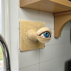 Könnte beinhalten: Keramik-Wandskulptur mit einem detaillierten Auge mit blauer Iris, montiert auf einer rissigen, beigefarbenen Fliese. Das Auge ragt aus der Fliese heraus. Die Skulptur ist an einer weiß gefliesten Wand mit einem Holzregal darüber befestigt.