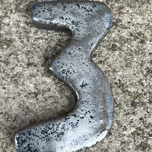 Op de afbeelding: Een zilverkleurig metalen nummer drie, waarschijnlijk voor een huis of bedrijf. Het nummer heeft een getextureerd, verweerd uiterlijk, wat suggereert dat het van metaal is gemaakt. Het nummer wordt weergegeven op een ruw, grijs betonnen oppervlak.