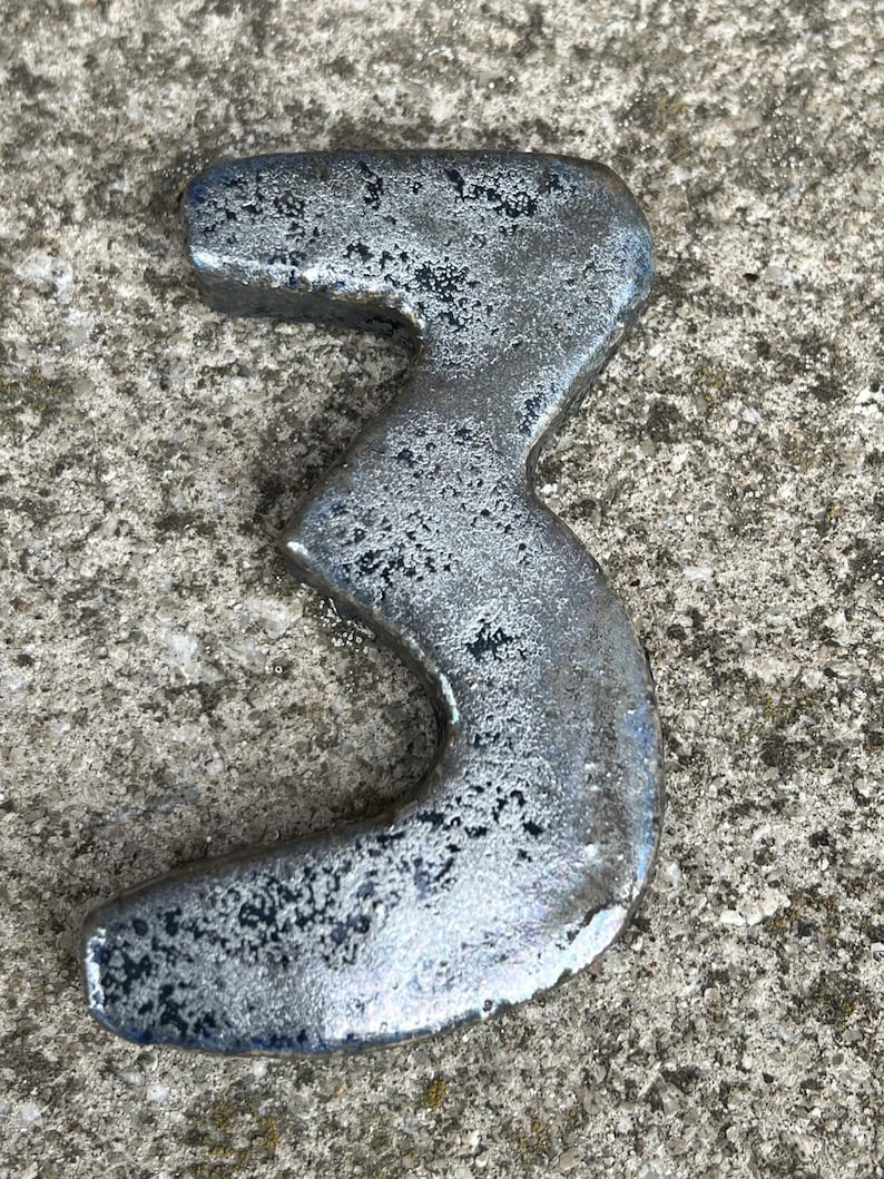 Op de afbeelding: Een zilverkleurig metalen nummer drie, waarschijnlijk voor een huis of bedrijf. Het nummer heeft een getextureerd, verweerd uiterlijk, wat suggereert dat het van metaal is gemaakt. Het nummer wordt weergegeven op een ruw, grijs betonnen oppervlak.