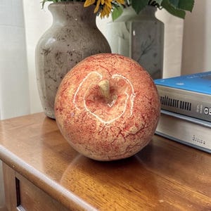 Op de afbeelding: Een decoratieve, ronde, rood-oranje appelsculptuur met een steel, op een houten tafel. Twee vazen met zonnebloemen en groen staan op de achtergrond. De appel heeft een gebarsten, getextureerd oppervlak.