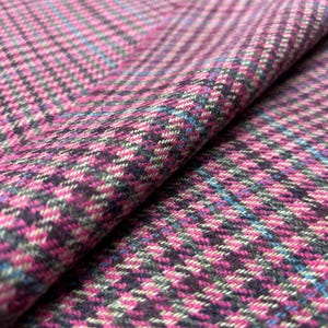 May include: Close-up of a folded fabric with a houndstooth pattern. The fabric features a blend of pink, black, blue, and gray threads, creating a textured appearance. The weave is visible, showcasing the detailed craftsmanship of the textile.