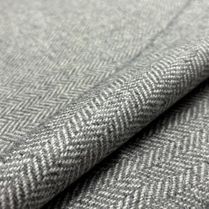 May include: Close-up of a gray and white herringbone fabric. The fabric is folded, revealing the texture and pattern. The weave creates a subtle, textured surface, suitable for apparel or home decor.