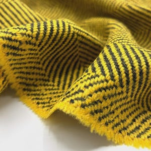 May include: Close-up of a yellow fabric with a black herringbone pattern. The fabric is folded, showing the texture and weave. The edges are frayed, indicating a cut piece of material. The color is a vibrant yellow with black stripes.