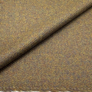 May include: Close-up of a folded fabric with a textured, speckled appearance. The fabric features a blend of brown, gold, and blue hues, creating a tweed-like effect. The edges of the fabric are visible, showing a fringed detail.