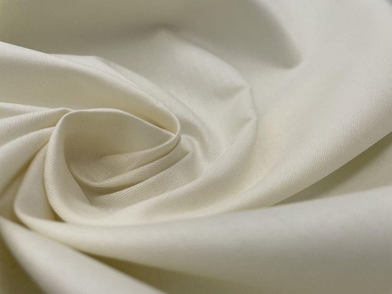 May include: Close-up of off-white fabric with soft folds and a subtle sheen. The material is tightly woven, suggesting a smooth texture. The fabric's color is a creamy, neutral tone, ideal for various sewing projects.