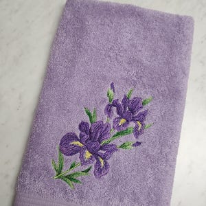 May include: A lavender-colored bath towel with an embroidered floral design. The embroidery features purple irises with green leaves and yellow accents. The towel is rectangular and has a textured surface.
