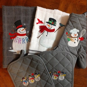 May include: Set of four gray kitchen towels with embroidered snowman designs. Two towels feature a single snowman, one towel features a snowman with a chef's hat, and one towel features three snowmen. The towels are quilted and have a diamond pattern.