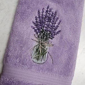 May include: A lavender-colored bath towel with an embroidered design of lavender sprigs in a mason jar tied with a ribbon. The towel has a textured surface and a decorative border.