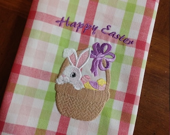 May include: A white kitchen towel with a pink, green, and white checkered pattern. The towel has an embroidered design of a bunny rabbit holding a basket of Easter eggs with the text "Happy Easter" above the bunny.