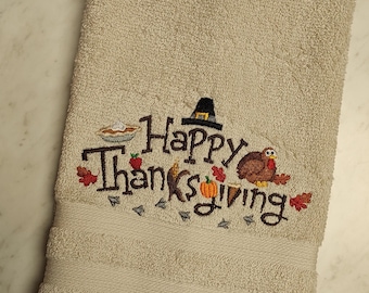 May include: A beige hand towel featuring embroidered text that reads Happy Thanksgiving. The design includes a black pilgrim hat, a brown turkey, a small pumpkin, a pie, and several red and orange autumn leaves stitched onto the fabric.