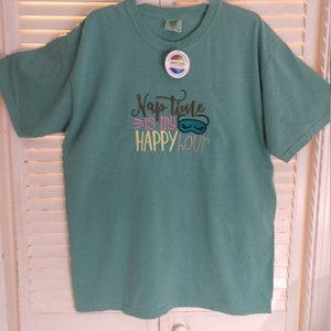May include: A teal blue t-shirt with the text "Nap time is my happy hour" embroidered on the front. The text is in a variety of colors, including pink, yellow, and green. A sleep mask is embroidered above the text.