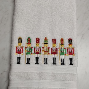 May include: White bath towel with a row of embroidered nutcracker figures in red, green, gold, and black.