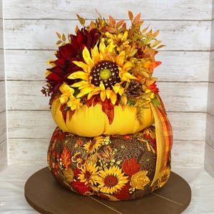 May include: A two-tiered decorative pumpkin arrangement. The top pumpkin is yellow and adorned with sunflowers, dahlias, and fall foliage. The bottom pumpkin is brown with a sunflower and leaf pattern. A decorative ribbon adds a touch of color.
