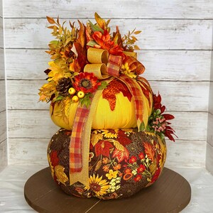 May include: A decorative, two-tiered pumpkin arrangement. The top pumpkin is yellow, adorned with a plaid ribbon and fall foliage. The bottom pumpkin is covered in a floral fabric with sunflowers and leaves. The arrangement sits on a wooden base.