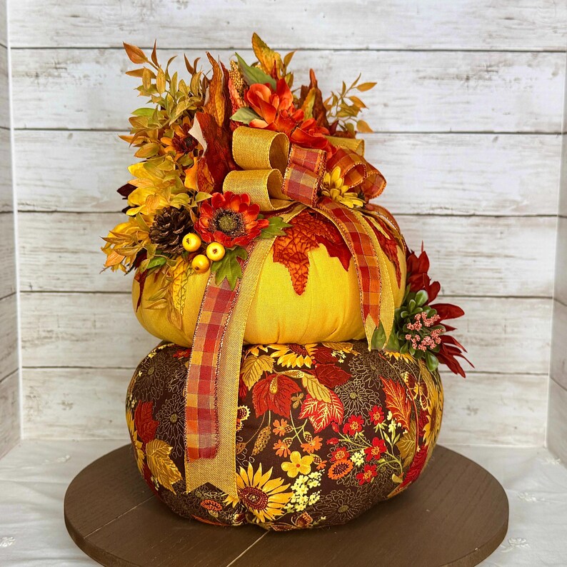 May include: A decorative, two-tiered pumpkin arrangement. The top pumpkin is yellow, adorned with a plaid ribbon and fall foliage. The bottom pumpkin is covered in a floral fabric with sunflowers and leaves. The arrangement sits on a wooden base.