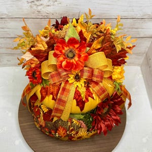 May include: A decorative fall-themed centerpiece featuring a pumpkin shape. The pumpkin is adorned with colorful autumn leaves, sunflowers, and a large orange flower. A golden yellow ribbon and a plaid ribbon add a festive touch. The centerpiece sits on a wooden base.