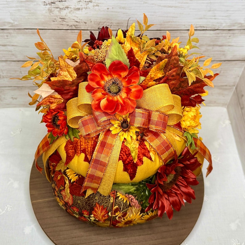 May include: A decorative fall-themed centerpiece featuring a pumpkin shape. The pumpkin is adorned with colorful autumn leaves, sunflowers, and a large orange flower. A golden yellow ribbon and a plaid ribbon add a festive touch. The centerpiece sits on a wooden base.