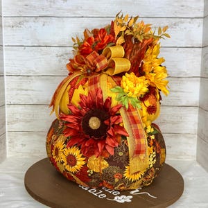 May include: A decorative, two-tiered pumpkin with a sunflower and floral design. The pumpkin is adorned with a large red sunflower, yellow and orange ribbons, and a variety of fall-colored flowers. It sits on a round wooden base.