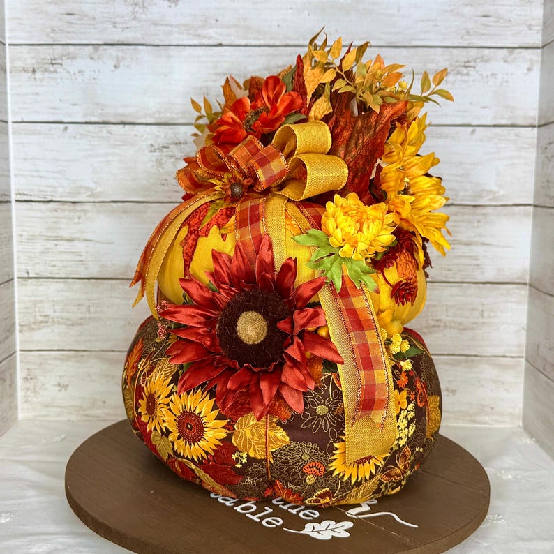 May include: A decorative, two-tiered pumpkin with a sunflower and floral design. The pumpkin is adorned with a large red sunflower, yellow and orange ribbons, and a variety of fall-colored flowers. It sits on a round wooden base.