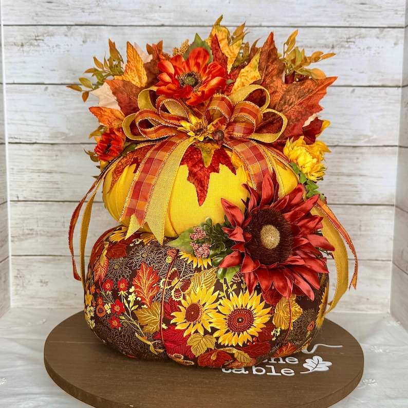 May include: A decorative, two-tiered pumpkin arrangement. The top pumpkin is yellow with a plaid ribbon and fall foliage. The bottom pumpkin is covered in a floral fabric with sunflowers and leaves. A large red sunflower and other flowers adorn the arrangement.