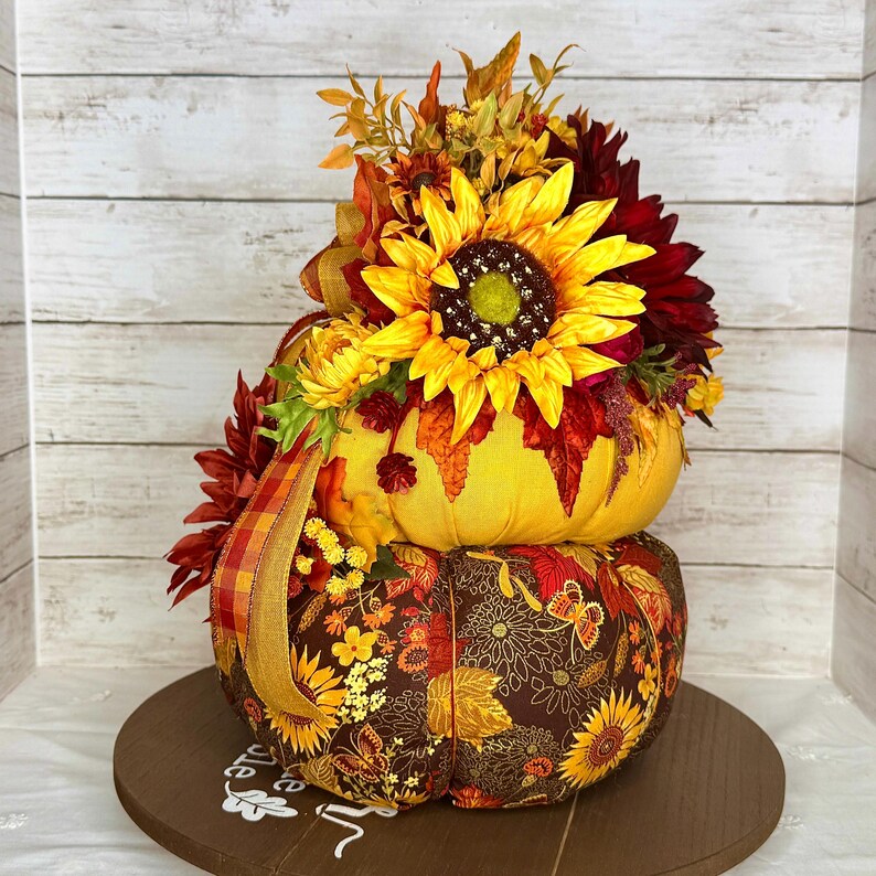 May include: A decorative, stacked pumpkin with a sunflower and fall foliage arrangement. The top pumpkin is yellow, and the bottom pumpkin is brown with a floral pattern. A wooden base supports the pumpkins, creating a festive autumn display.