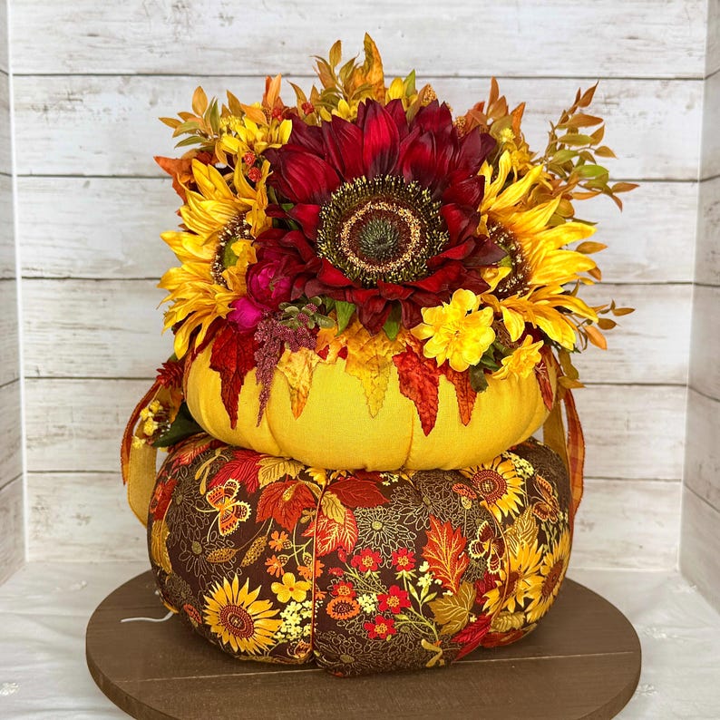 May include: A decorative, two-tiered pumpkin arrangement. The top pumpkin is yellow and adorned with a variety of sunflowers and fall foliage. The bottom pumpkin is covered in a brown fabric with a floral and leaf pattern. The arrangement sits on a wooden base.