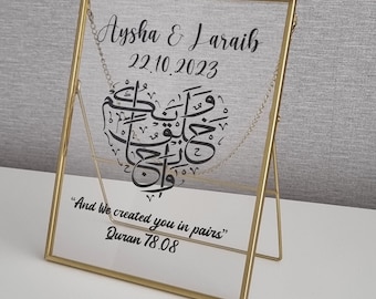 May include: A gold-framed glass picture with a white background. The picture features a black Arabic calligraphy design in the shape of a heart. The text "Aysha & Laraib 22.10.2023" is at the top of the picture. The text "And We created you in pairs" Quran 78.08 is at the bottom of the picture.