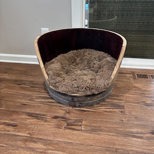 May include: A wooden dog bed made from a repurposed wine barrel. The bed has a brown faux fur liner and is sitting on a hardwood floor.