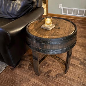 May include: A round wooden table made from a repurposed barrel with a black metal band and a black metal X-shaped base. The table is 24 inches in diameter and 20 inches tall. A glass with a drink is on the table.