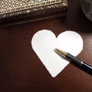 May include: A white paper heart shape on a brown wooden surface with a black and gold pen lying on top of it.