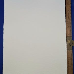 May include: A rectangular, off-white watercolor paper sheet with a textured surface. A wooden ruler with inch markings is positioned along the right edge. The paper is on a blue surface.