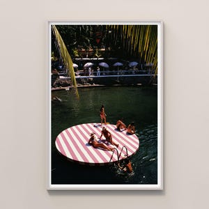 Peut inclure: Une impression encadrée présentant une photographie vintage d'une scène de piscine. Une plateforme ronde à rayures roses et blanches flotte dans l'eau, avec des personnes prenant le soleil et nageant. Des palmiers et des parasols sont visibles en arrière-plan.