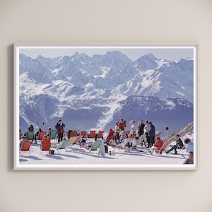 Könnte beinhalten: Ein gerahmter Druck mit einer Vintage-Skigebietszene. Menschen entspannen sich in bunten Liegestühlen auf einer schneebedeckten Piste, mit schneebedeckten Bergen im Hintergrund. Der Druck hat einen weißen Rand.