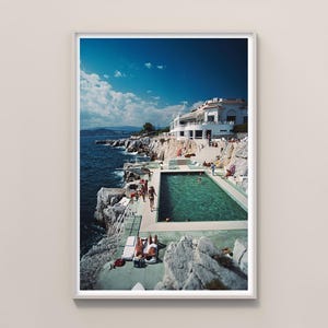 May include: A framed print of a coastal scene featuring a turquoise pool, white buildings, and the deep blue sea under a partly cloudy sky. People are seen sunbathing and swimming, evoking a sense of leisure and vacation.