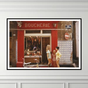 May include: A framed photograph of a French butcher shop, with the word "BOUCHERIE" above the entrance. Three women stand in front of the shop. A bicycle is parked in front of the shop. The shop has red trim and a tiled wall.