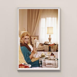 May include: A framed vintage photograph of a woman in a teal dress and fur collar reading a newspaper in a bedroom. A tray with food and a teacup sits on the bed. A lamp and curtains are visible in the background.