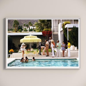 May include: A framed print of a vintage outdoor pool party scene. A yellow umbrella shades a table with guests in stylish attire. People are in the pool, and others are standing around. The setting is a mid-century modern home with lush landscaping.