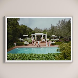Könnte beinhalten: Gerahmtes Foto einer Poolparty-Szene. Ein türkisfarbener Pool ist von Grün und einer Backstein-Terrasse umgeben. Menschen versammeln sich in der Nähe eines weißen Pavillons und um den Pool, mit weißen Sonnenschirmen und Blumengestecken.