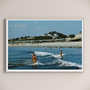 Può includere: Una stampa incorniciata di una scena di spiaggia vintage. I surfisti cavalcano le onde in primo piano, con edifici e persone sulla riva sullo sfondo. Il cielo è blu con nuvole sparse, creando un'atmosfera estiva nostalgica.