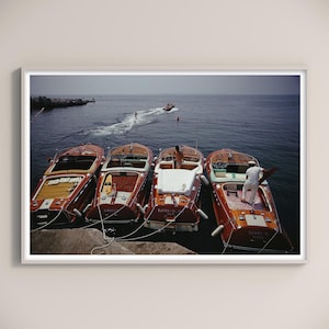 May include: A framed print featuring four classic wooden motorboats docked at a pier. The boats are a rich reddish-brown with white accents. A person stands in one boat, and another boat is seen in the distance on the water.