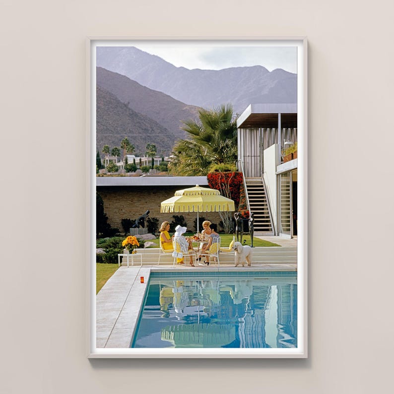 May include: A framed print of a vintage scene featuring a pool, patio, and mountain backdrop. Three people are seated at a table under a yellow umbrella. A white dog stands nearby. The pool's water reflects the sky and surrounding elements.