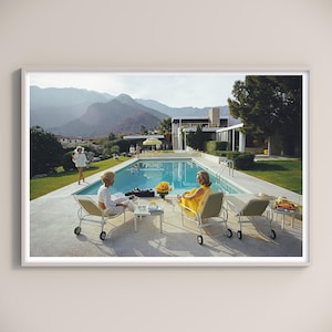 May include: A framed print showing a mid-century scene with a swimming pool, sun loungers, and people. Two women relax by the pool, one in a yellow dress. Mountains and a large house are in the background. The print is in a white frame.