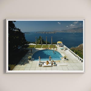 May include: A framed print of a vintage scene featuring a turquoise pool, ocean, and blue sky. People are sunbathing and swimming. The scene includes white pillars, a yellow umbrella, and a white terrace.