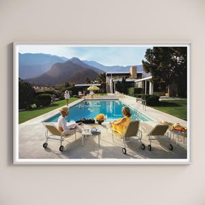 May include: A framed print depicting a retro scene by a swimming pool. Two women in patterned and yellow dresses relax on lounge chairs. A pool, mountains, and a mid-century modern house are in the background. Other people are visible near the pool.