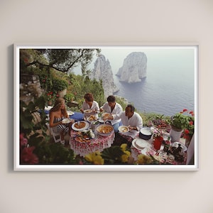 Puede incluir: Fotografía enmarcada que muestra a un grupo de personas cenando al aire libre. Están sentados a mesas repletas de comida, con vistas a una vista costera con grandes formaciones rocosas en el mar. La escena está bañada por la luz natural.
