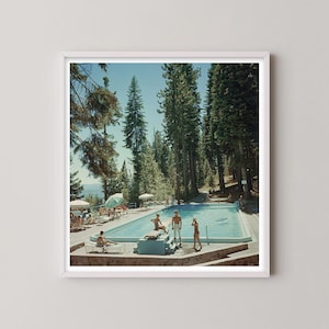 May include: A framed print of a vintage photograph featuring a swimming pool scene. People are lounging by the pool and in the water. Tall pine trees surround the pool, and the sky is a clear blue. The print is in a white frame.